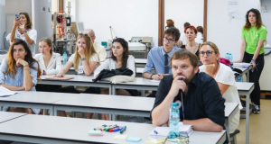 A room with white desks where women and men, aged between 20 and 40, look to the same direction, to the front.